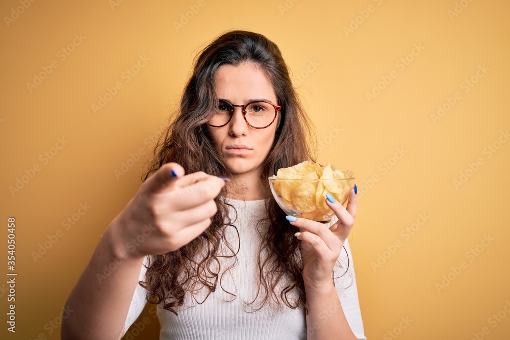 Young beautiful woman with curly hair holding bowl of chips potatoes over yellow background pointing with finger to the camera and to you, hand sign, positive and confident gesture from the front