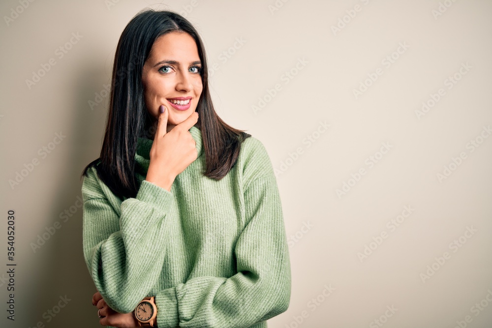 Young brunette woman with blue eyes wearing turtleneck sweater over white background looking confident at the camera smiling with crossed arms and hand raised on chin. Thinking positive. Young brunette woman with blue eyes wearing turtleneck sweater over white background looking confident at the camera smiling with crossed arms and hand raised on chin. Thinking positive.