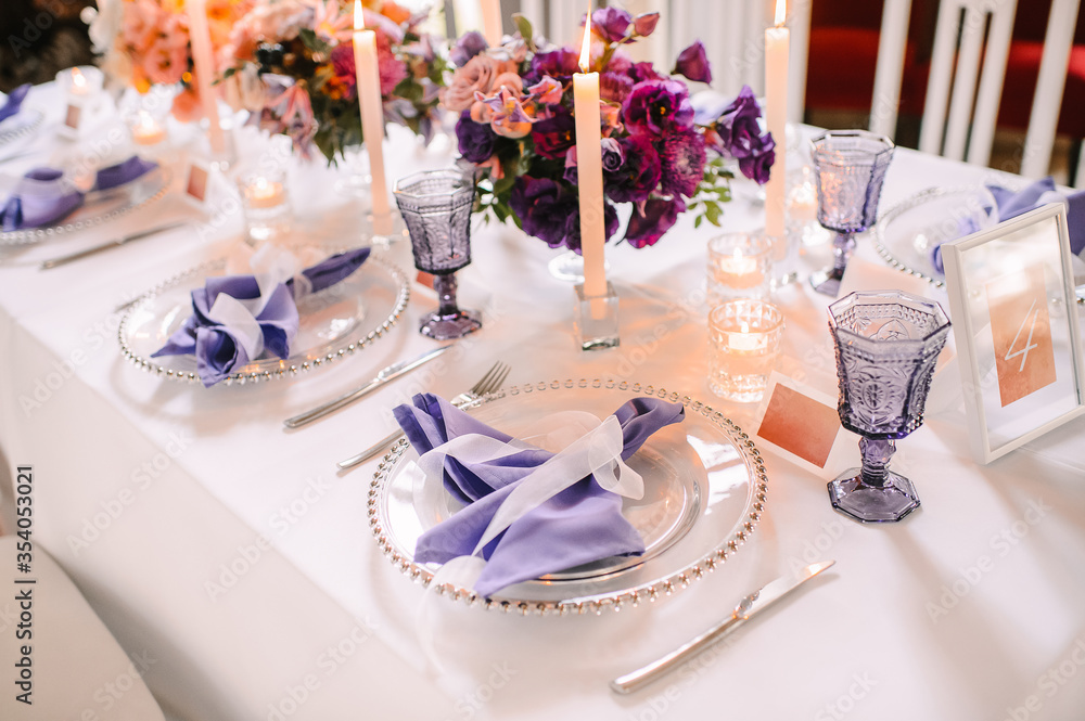 Fototapeta premium Presidium, banquet table with a white tablecloth. Plates and cutlery, purple glasses, napkins, candles in glass candlesticks. Bouquet of flowers, asters, roses, carnations, eustomas.
