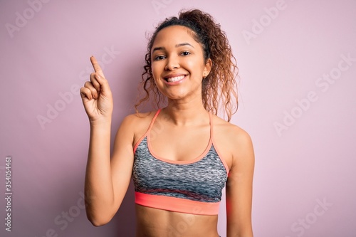 Beautiful sportswoman with curly hair doing sport wearing sportswear over pink background showing and pointing up with finger number one while smiling confident and happy.