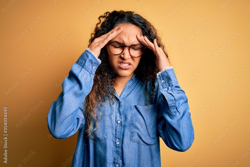 Beautiful woman with curly hair wearing casual denim shirt and glasses over yellow background with hand on head for pain in head because stress. Suffering migraine. Beautiful woman with curly hair wearing casual denim shirt and glasses over yellow background with hand on head for pain in head because stress. Suffering migraine.