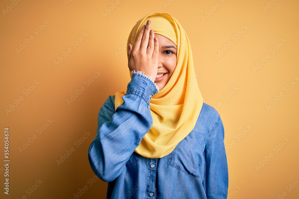 Young beautiful woman with curly hair wearing arab traditional hijab over yellow background covering one eye with hand, confident smile on face and surprise emotion. Young beautiful woman with curly hair wearing arab traditional hijab over yellow background covering one eye with hand, confident smile on face and surprise emotion.