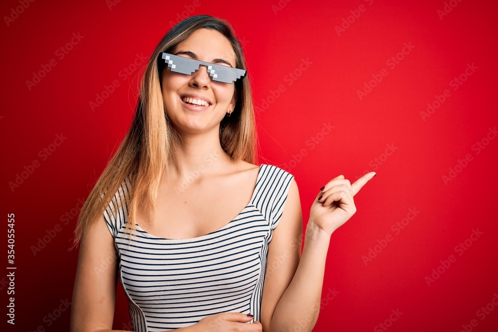 Young beautiful brunette woman wearing funny thug life sunglasses over red background with a big smile on face, pointing with hand and finger to the side looking at the camera. Young beautiful brunette woman wearing funny thug life sunglasses over red background with a big smile on face, pointing with hand and finger to the side looking at the camera.