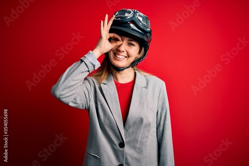 Young beautiful blonde motorcyclist woman wearing motorcycle helmet over red background doing ok gesture with hand smiling, eye looking through fingers with happy face. © Krakenimages.com