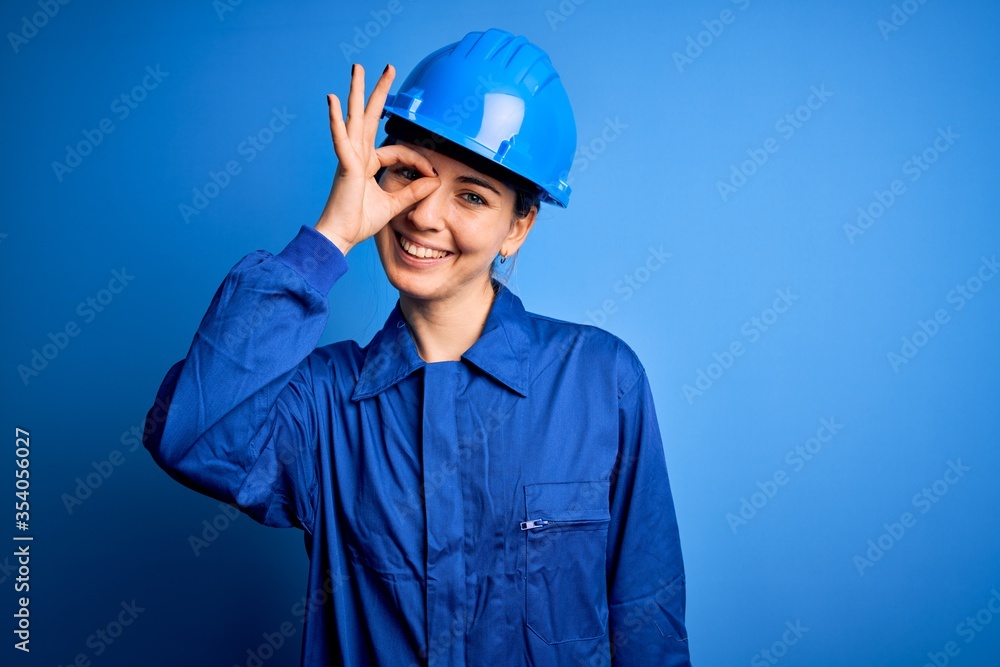 Young beautiful worker woman with blue eyes wearing security helmet and uniform doing ok gesture with hand smiling, eye looking through fingers with happy face. Young beautiful worker woman with blue eyes wearing security helmet and uniform doing ok gesture with hand smiling, eye looking through fingers with happy face.
