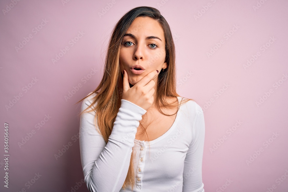 Young beautiful blonde woman with blue eyes wearing white t-shirt over pink background Looking fascinated with disbelief, surprise and amazed expression with hands on chin Young beautiful blonde woman with blue eyes wearing white t-shirt over pink background Looking fascinated with disbelief, surprise and amazed expression with hands on chin