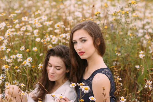 Two girls in dark blue and white dresses in sunny day sitting in chamomile field