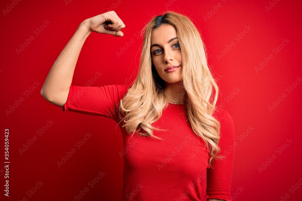Naklejka premium Young beautiful blonde woman wearing casual t-shirt standing over isolated red background Strong person showing arm muscle, confident and proud of power