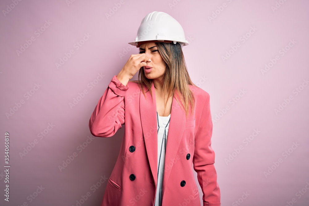 Young beautiful brunette architect woman wearing safety helmet over pink background smelling something stinky and disgusting, intolerable smell, holding breath with fingers on nose. Bad smell Young beautiful brunette architect woman wearing safety helmet over pink background smelling something stinky and disgusting, intolerable smell, holding breath with fingers on nose. Bad smell