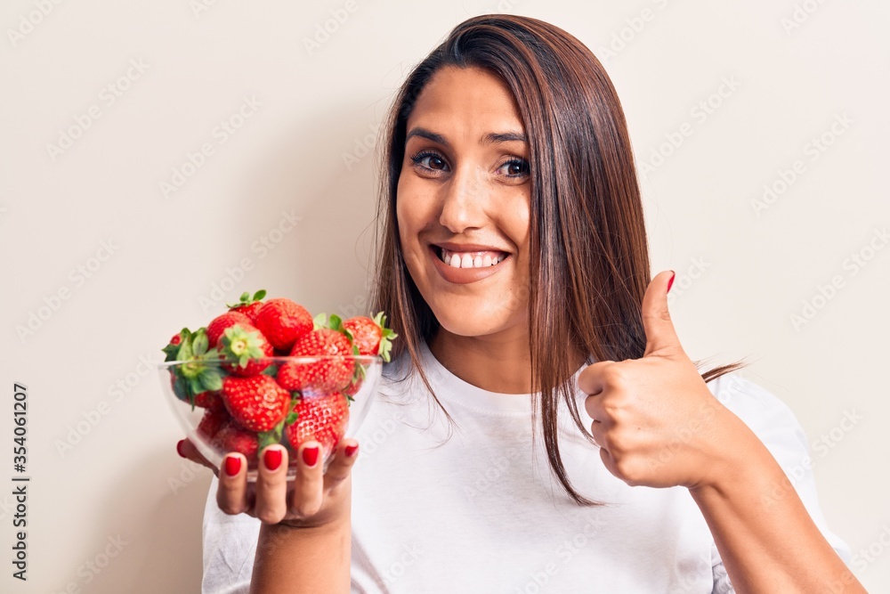 Young beautiful brunette woman holding bowl with strawberries smiling happy and positive, thumb up doing excellent and approval sign Young beautiful brunette woman holding bowl with strawberries smiling happy and positive, thumb up doing excellent and approval sign
