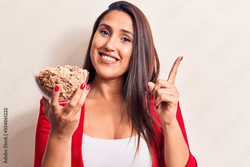 Young beautiful brunette woman holding bowl of cornflakes cereal smiling happy pointing with hand and finger to the side Young beautiful brunette woman holding bowl of cornflakes cereal smiling happy pointing with hand and finger to the side
