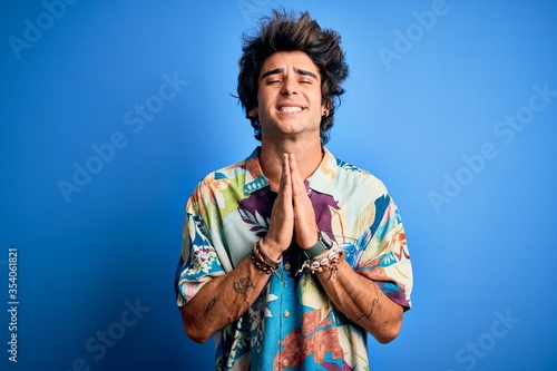 Young handsome man on vacation wearing summer shirt over isolated blue background begging and praying with hands together with hope expression on face very emotional and worried. Begging.
