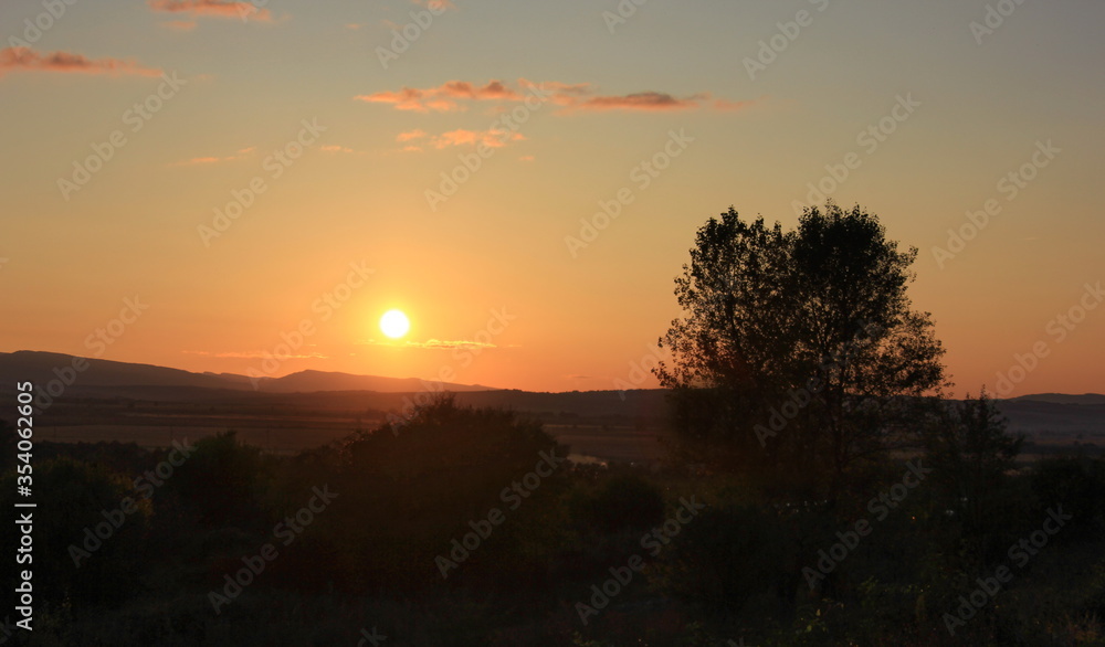 Sunset, summer landscape. Against the background of a peach-colored sky, the disk of the setting sun and the silhouettes of the woods and mountains in the background