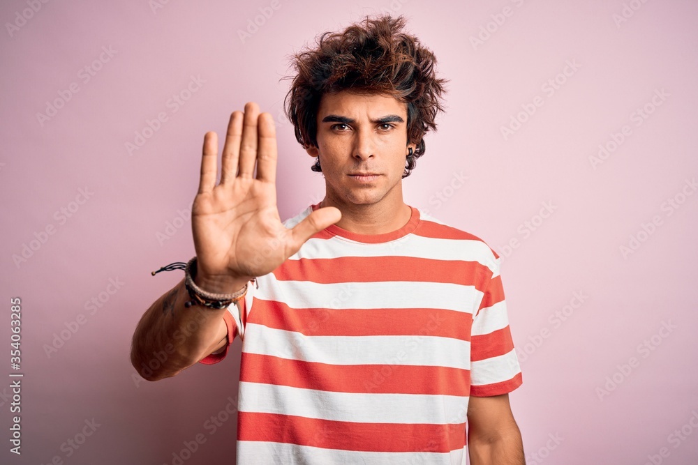 Young handsome man wearing striped casual t-shirt standing over isolated pink background doing stop sing with palm of the hand. Warning expression with negative and serious gesture on the face. Young handsome man wearing striped casual t-shirt standing over isolated pink background doing stop sing with palm of the hand. Warning expression with negative and serious gesture on the face.