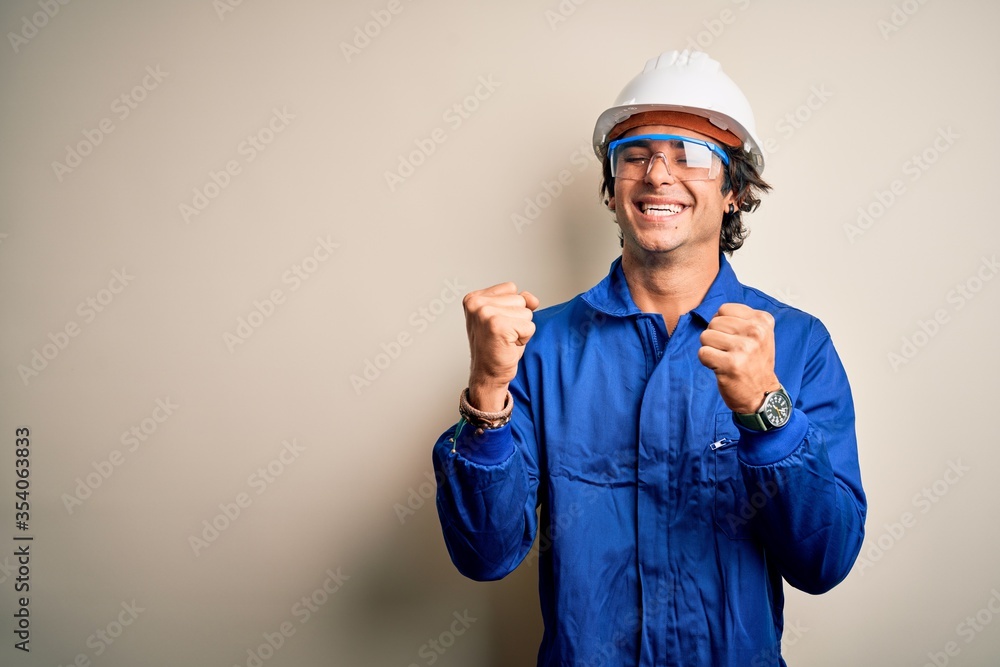 Young constructor man wearing uniform and security helmet over isolated white background very happy and excited doing winner gesture with arms raised, smiling and screaming for success. Celebration