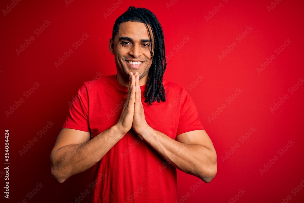 Young handsome african american afro man with dreadlocks wearing red casual t-shirt praying with hands together asking for forgiveness smiling confident. Young handsome african american afro man with dreadlocks wearing red casual t-shirt praying with hands together asking for forgiveness smiling confident.