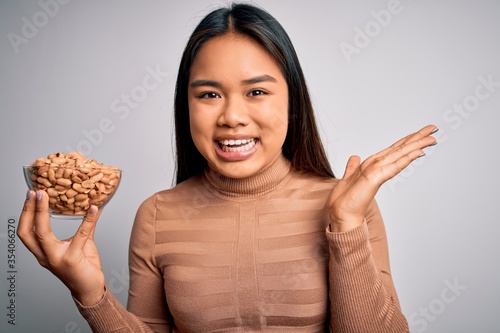 Young asian girl holding bowl of healthy peanuts eating as snack for diet over white background very happy and excited, winner expression celebrating victory screaming with big smile and raised hands
