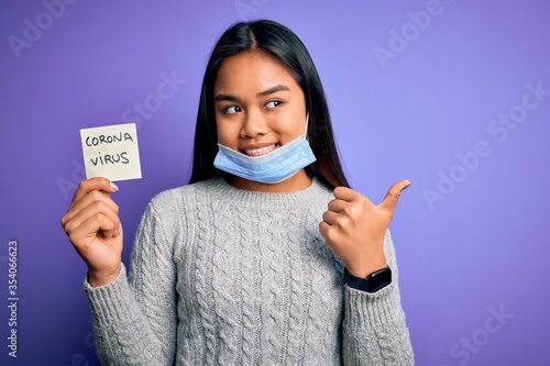 Asian girl wearing medical mask asking for alert holding reminder with corona virus message pointing and showing with thumb up to the side with happy face smiling