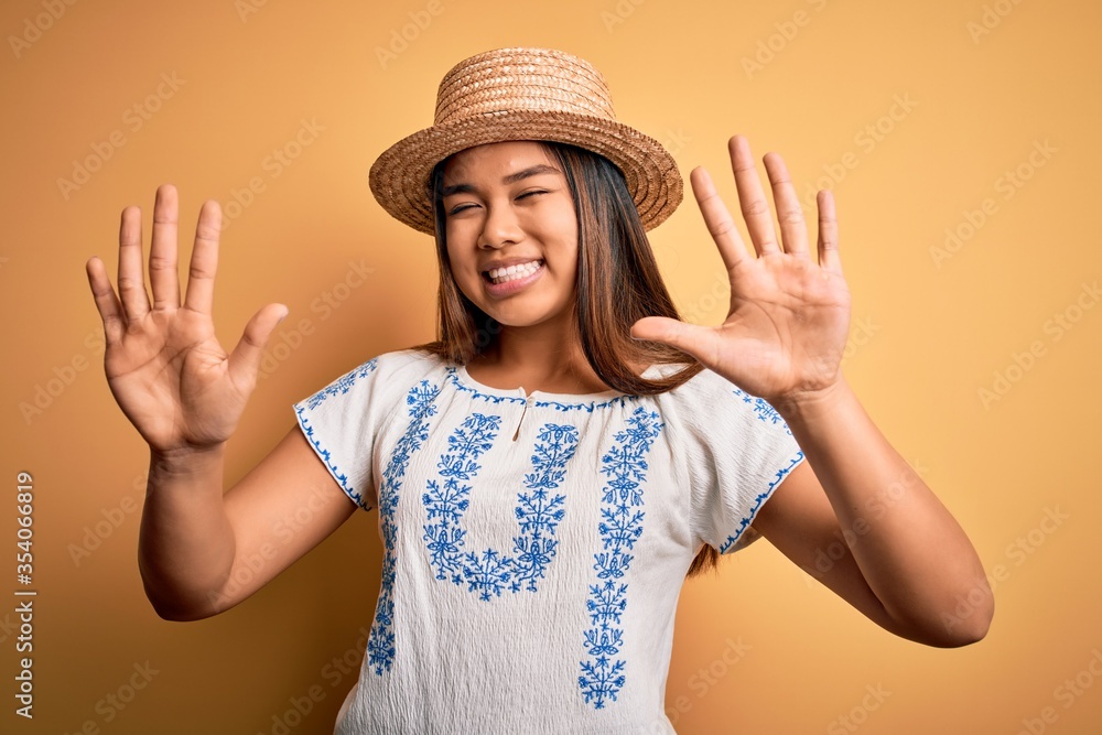 Young beautiful asian girl wearing casual t-shirt and hat standing over yellow background showing and pointing up with fingers number ten while smiling confident and happy. Young beautiful asian girl wearing casual t-shirt and hat standing over yellow background showing and pointing up with fingers number ten while smiling confident and happy.