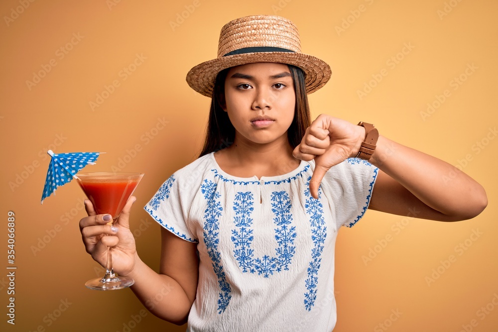 Young asian girl on vacation wearing summer hat drinking cocktail over yellow background with angry face, negative sign showing dislike with thumbs down, rejection concept Young asian girl on vacation wearing summer hat drinking cocktail over yellow background with angry face, negative sign showing dislike with thumbs down, rejection concept