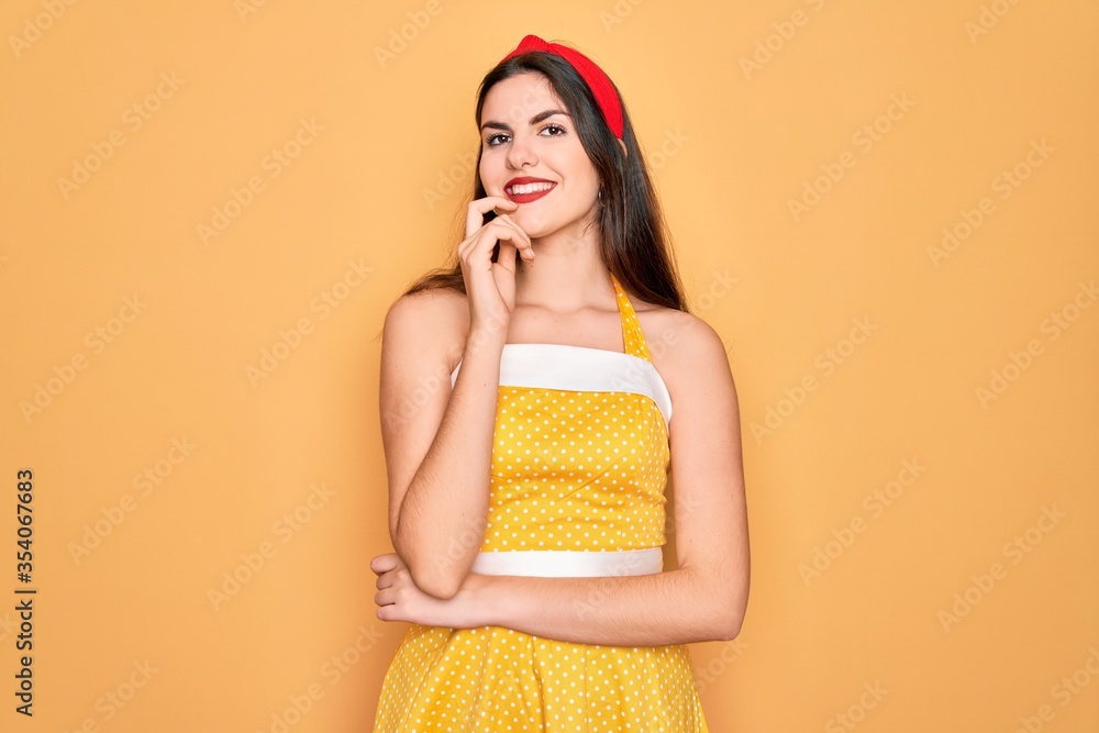 Young beautiful pin up woman wearing 50s fashion vintage dress over yellow background looking confident at the camera with smile with crossed arms and hand raised on chin. Thinking positive. Young beautiful pin up woman wearing 50s fashion vintage dress over yellow background looking confident at the camera with smile with crossed arms and hand raised on chin. Thinking positive.