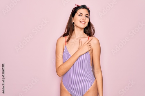Young beautiful fashion girl wearing swimwear swimsuit and sunglasses over pink background cheerful with a smile on face pointing with hand and finger up to the side with happy and natural expression