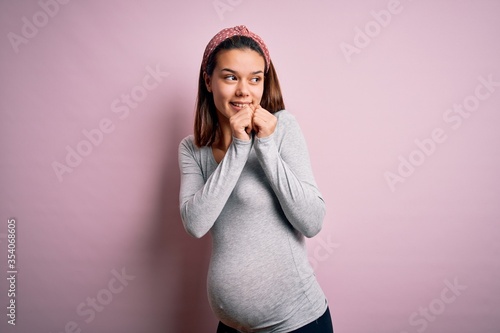 Young beautiful teenager girl pregnant expecting baby over isolated pink background laughing nervous and excited with hands on chin looking to the side