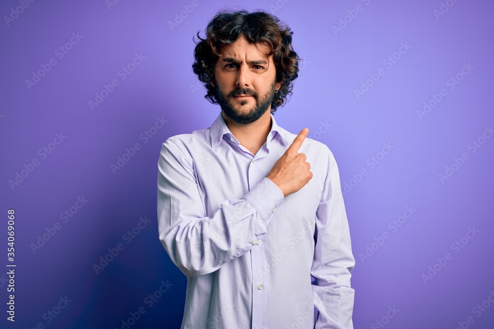 Young handsome business man with beard wearing shirt standing over purple background Pointing with hand finger to the side showing advertisement, serious and calm face Young handsome business man with beard wearing shirt standing over purple background Pointing with hand finger to the side showing advertisement, serious and calm face