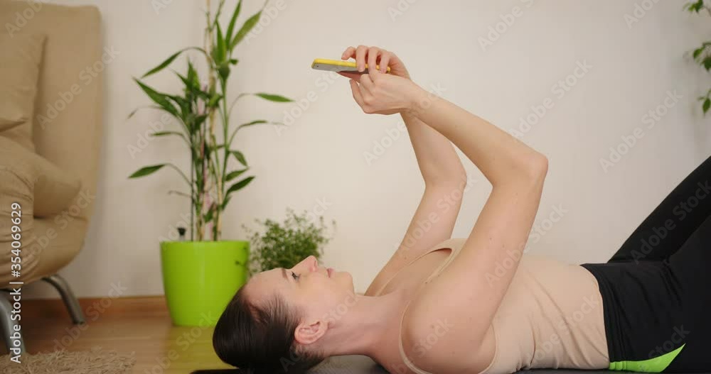 Athletic woman using smartphone while lying on yoga fitness mat at home. Athletic woman using smartphone while lying on yoga fitness mat at home.