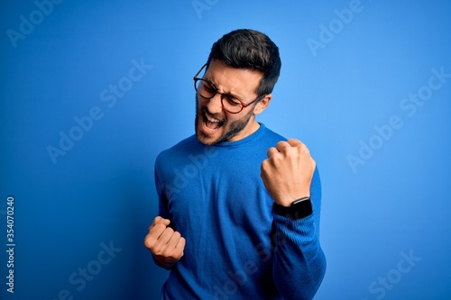 Photos Young handsome man with beard wearing casual sweater and glasses over blue background celebrating surprised and amazed for success with arms raised and eyes closed