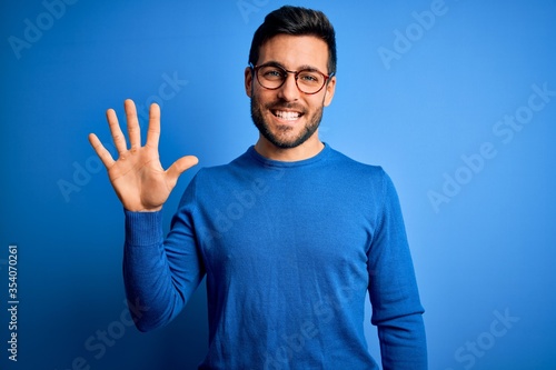 Foto Young handsome man with beard wearing casual sweater and glasses over blue background showing and pointing up with fingers number five while smiling confident and happy