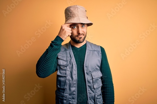 Handsome tourist man with beard on vacation wearing explorer hat over yellow background pointing unhappy to pimple on forehead, ugly infection of blackhead. Acne and skin problem