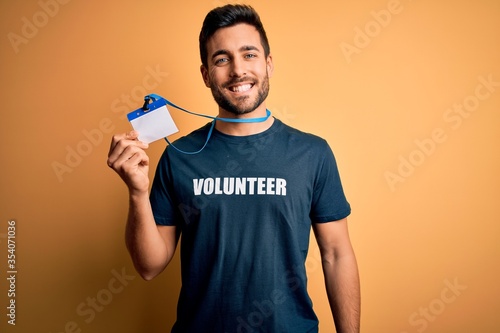 Handsome volunteer man with beard holding id card identification over yellow background with a happy face standing and smiling with a confident smile showing teeth © Krakenimages.com