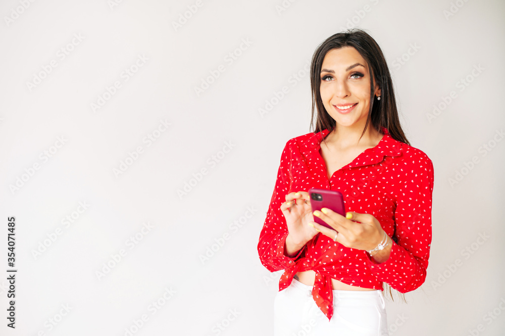 Beautiful young woman with a smartphone on a white background. Girl in red is typing on phone screen. Mockup, copyspace for text Beautiful young woman with a smartphone on a white background. Girl in red is typing on phone screen. Mockup, copyspace for text