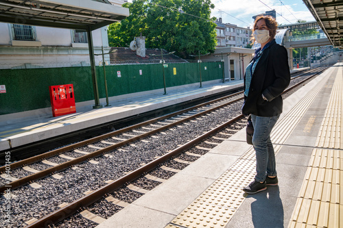 Young Caucasian casual dressed woman is waiting alone for the metro train at railway station. She wears protective medical face mask when using public transportation