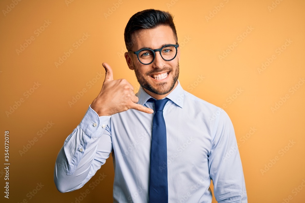 Young handsome businessman wearing tie and glasses standing over yellow background smiling doing phone gesture with hand and fingers like talking on the telephone. Communicating concepts. Young handsome businessman wearing tie and glasses standing over yellow background smiling doing phone gesture with hand and fingers like talking on the telephone. Communicating concepts.