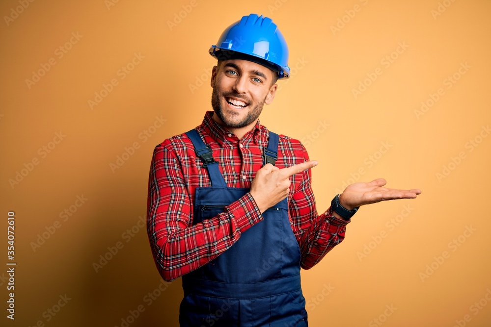 Young builder man wearing construction uniform and safety helmet over yellow background amazed and smiling to the camera while presenting with hand and pointing with finger.