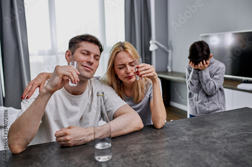 unscrupulous parents drink alcohol together at home while their child cry from hopelessness in the background.