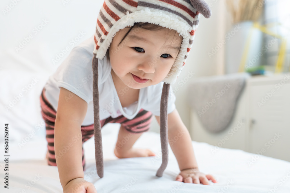 Asian little young cute baby kid lying and crawling on white bed in bedroom at home. The little child wearing bear hat playing on bed with happiness. Young baby activity and development growth concept Asian little young cute baby kid lying and crawling on white bed in bedroom at home. The little child wearing bear hat playing on bed with happiness. Young baby activity and development growth concept