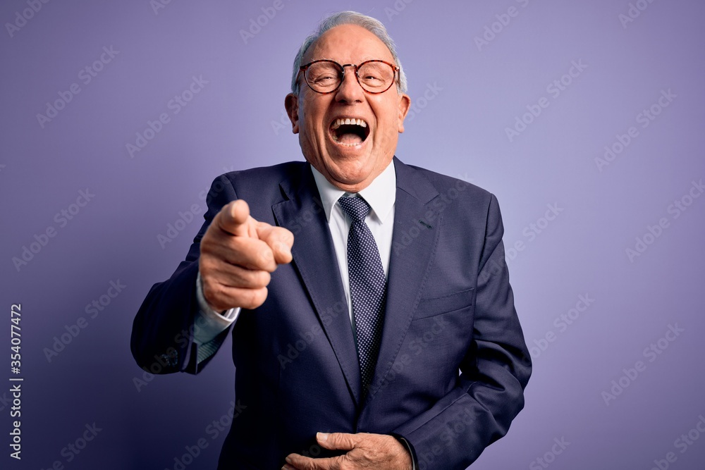 Grey haired senior business man wearing glasses and elegant suit and tie over purple background laughing at you, pointing finger to the camera with hand over body, shame expression Grey haired senior business man wearing glasses and elegant suit and tie over purple background laughing at you, pointing finger to the camera with hand over body, shame expression