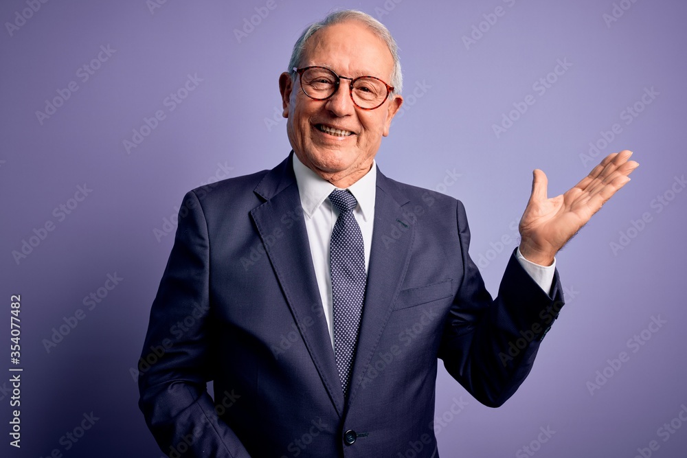 Grey haired senior business man wearing glasses and elegant suit and tie over purple background smiling cheerful presenting and pointing with palm of hand looking at the camera. Grey haired senior business man wearing glasses and elegant suit and tie over purple background smiling cheerful presenting and pointing with palm of hand looking at the camera.