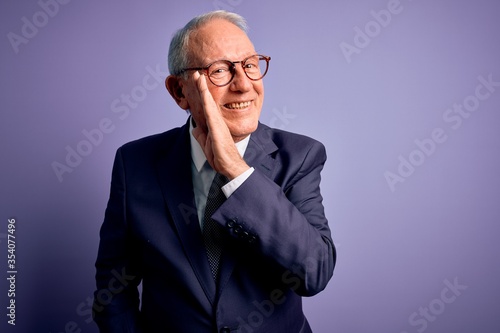 Grey haired senior business man wearing glasses and elegant suit and tie over purple background hand on mouth telling secret rumor, whispering malicious talk conversation