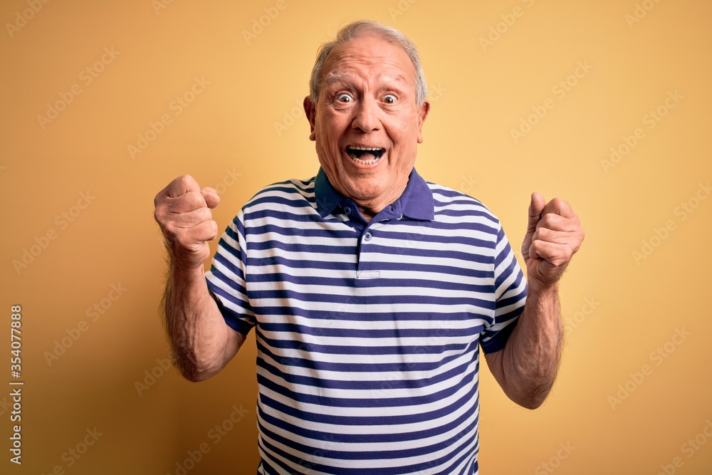 Grey haired senior man wearing casual navy striped t-shirt standing over yellow background celebrating surprised and amazed for success with arms raised and open eyes. Winner concept. Grey haired senior man wearing casual navy striped t-shirt standing over yellow background celebrating surprised and amazed for success with arms raised and open eyes. Winner concept.