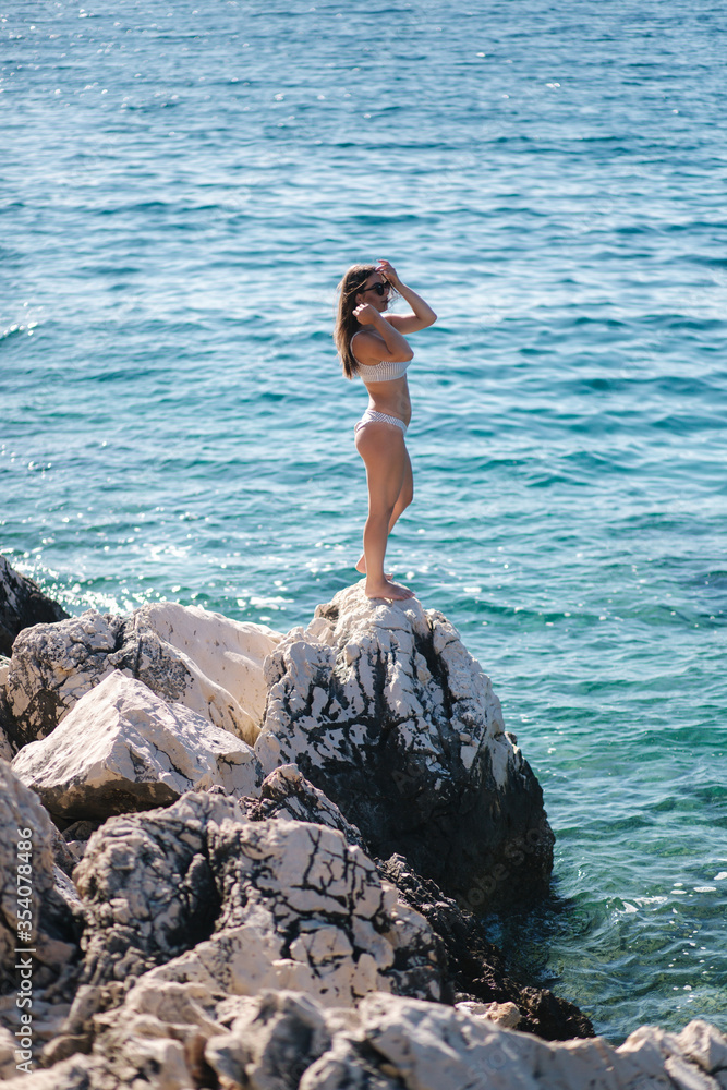 Sexy woman in bikini stand on the rock by the sea. Beautiful lagoon. Woman enjoying summer sun. Back view of sexy woman in bikini Sexy woman in bikini stand on the rock by the sea. Beautiful lagoon. Woman enjoying summer sun. Back view of sexy woman in bikini