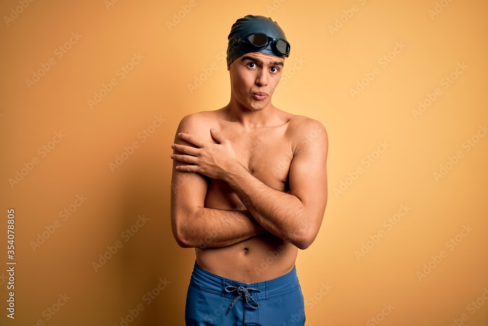 Young handsome man shirtless wearing swimsuit and swim cap over isolated yellow background shaking and freezing for winter cold with sad and shock expression on face