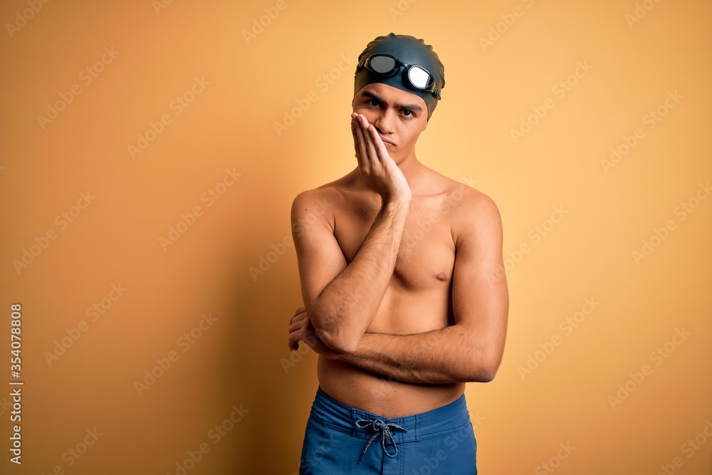 Young handsome man shirtless wearing swimsuit and swim cap over isolated yellow background thinking looking tired and bored with depression problems with crossed arms.