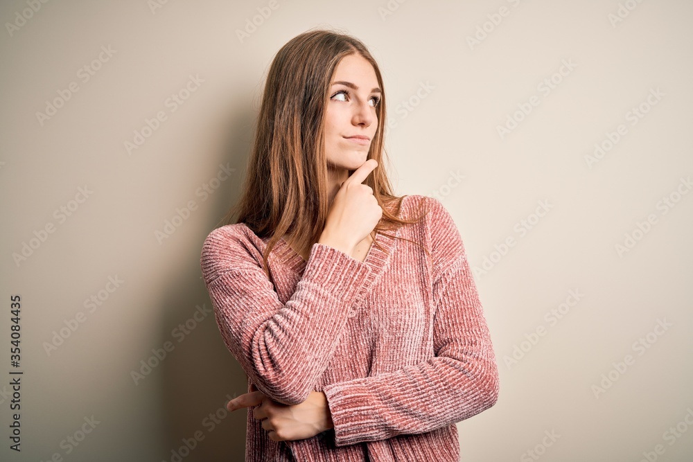 Young beautiful redhead woman wearing pink casual sweater over isolated white background with hand on chin thinking about question, pensive expression. Smiling with thoughtful face. Doubt concept. Young beautiful redhead woman wearing pink casual sweater over isolated white background with hand on chin thinking about question, pensive expression. Smiling with thoughtful face. Doubt concept.