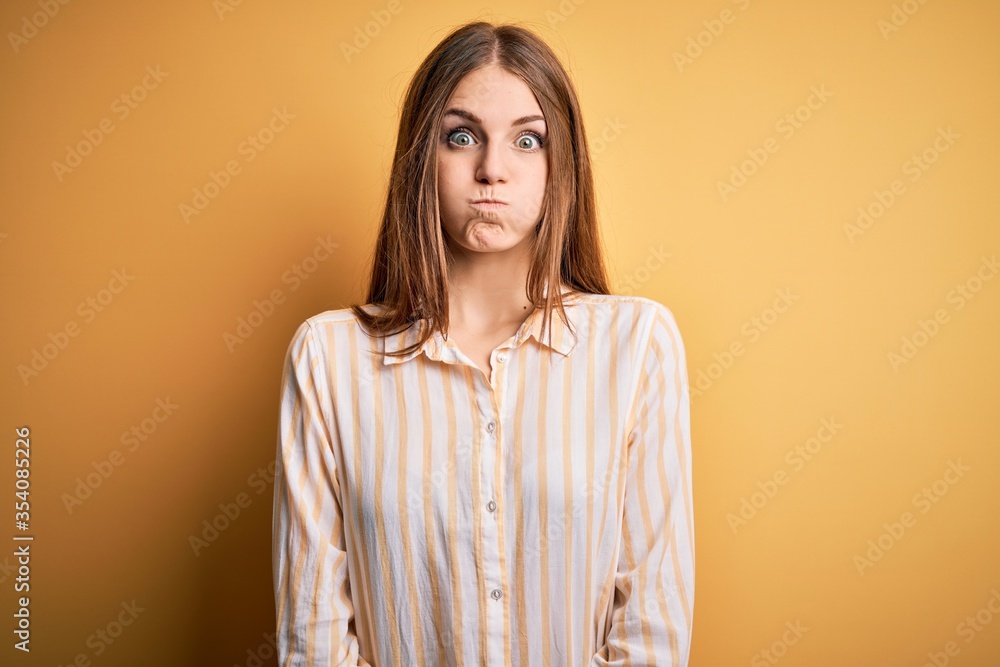 Young beautiful redhead woman wearing casual striped shirt over isolated yellow background puffing cheeks with funny face. Mouth inflated with air, crazy expression. Young beautiful redhead woman wearing casual striped shirt over isolated yellow background puffing cheeks with funny face. Mouth inflated with air, crazy expression.