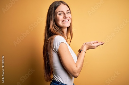 Young beautiful redhead woman wearing casual t-shirt over isolated yellow background pointing aside with hands open palms showing copy space, presenting advertisement smiling excited happy