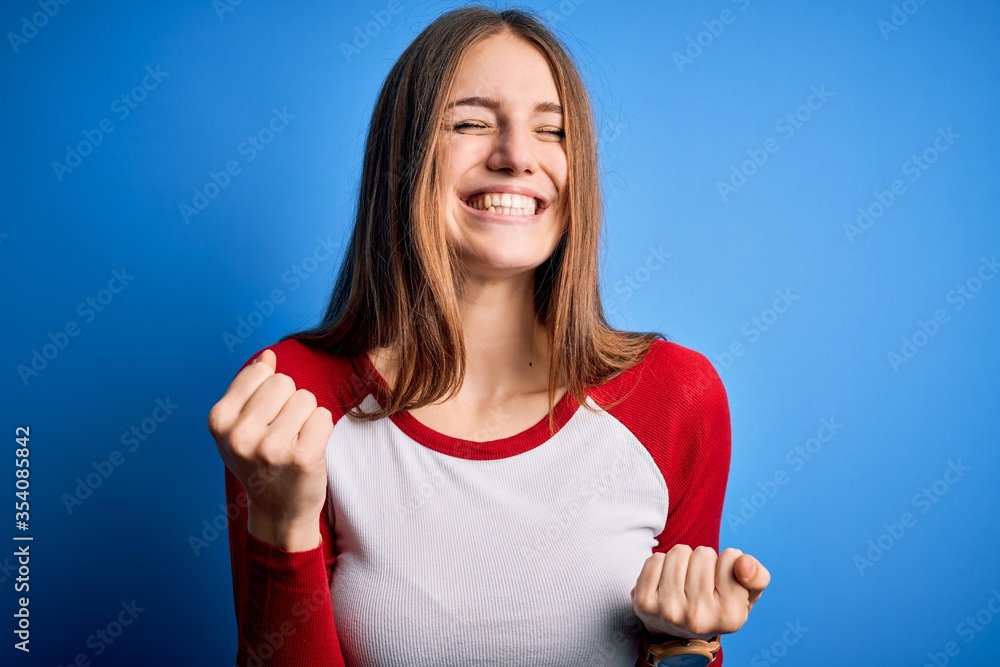 Young beautiful redhead woman wearing casual t-shirt over isolated blue background very happy and excited doing winner gesture with arms raised, smiling and screaming for success. Celebration concept. Young beautiful redhead woman wearing casual t-shirt over isolated blue background very happy and excited doing winner gesture with arms raised, smiling and screaming for success. Celebration concept.